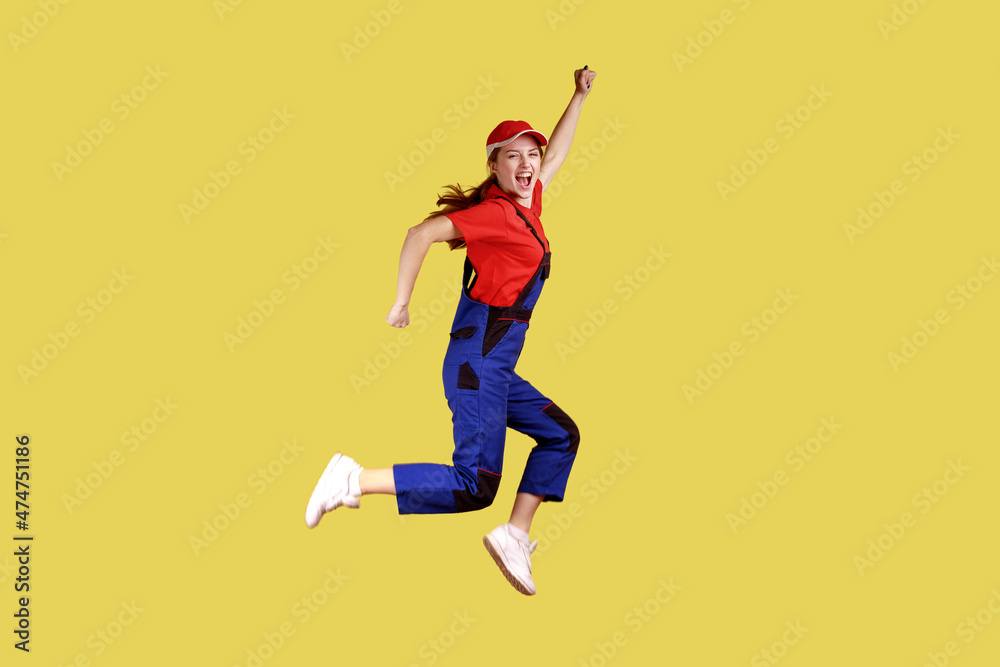 Side view portrait of extremely happy worker woman jumping and clenched fists, celebrating long awaited day off, wearing overalls and red cap. Indoor studio shot isolated on yellow background. Side view portrait of extremely happy worker woman jumping and clenched fists, celebrating long awaited day off, wearing overalls and red cap. Indoor studio shot isolated on yellow background.