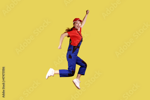 Side view portrait of extremely happy worker woman jumping and clenched fists, celebrating long awaited day off, wearing overalls and red cap. Indoor studio shot isolated on yellow background.