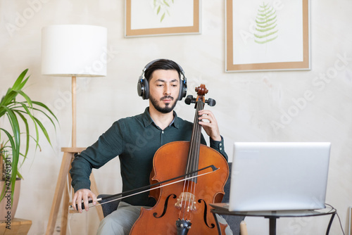 Young confident teacher of music in headphones playing string musical instrument during online lesson
