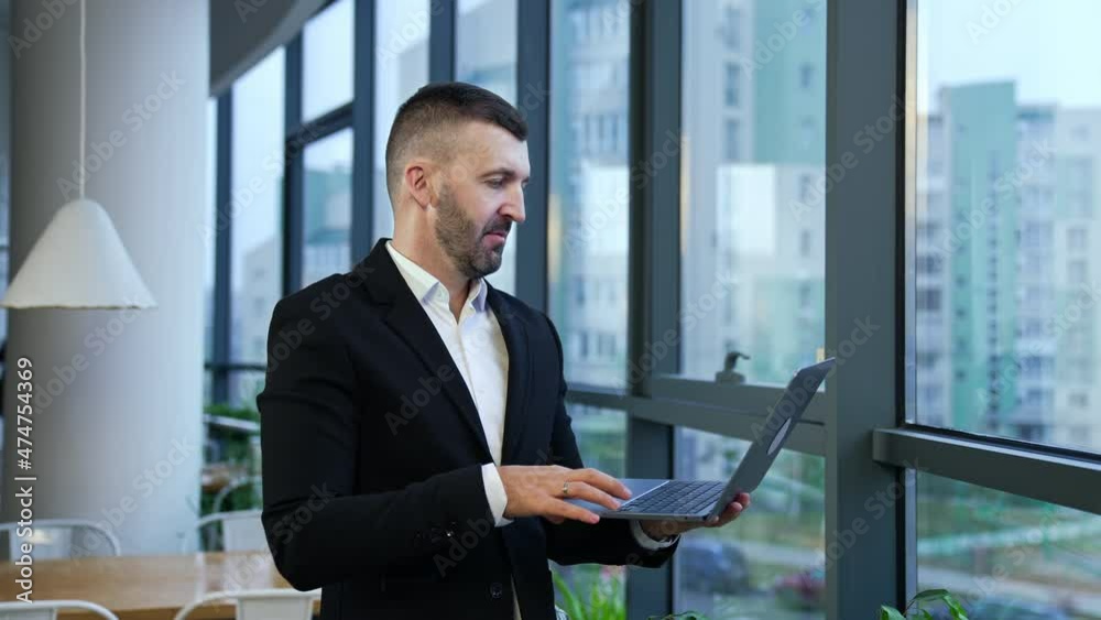 Male entrepreneur standing at the window and holding laptop in front of him. Business talk on the computer. Man talking and gesturing in front of notebook. Male entrepreneur standing at the window and holding laptop in front of him. Business talk on the computer. Man talking and gesturing in front of notebook.