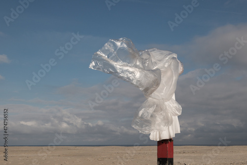 Abstract portrait of woman in white dress and palstic sheet on beach pole near clouds in wind photo