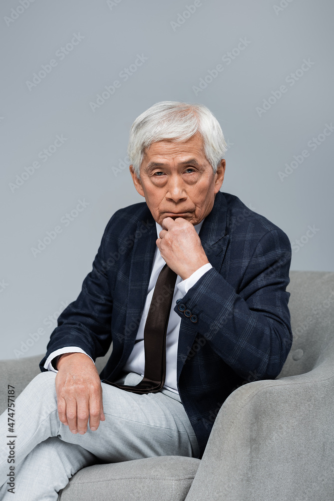 senior asian businessman in formal wear looking at camera while sitting in armchair isolated on grey senior asian businessman in formal wear looking at camera while sitting in armchair isolated on grey