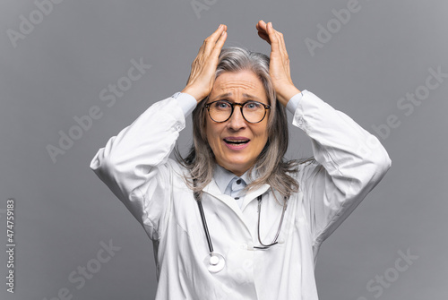 Senior doctor in white medical gown with stethoscope on shoulders staring at the camera with fear on the face, grab head isolated on gray, mature physical therapist feels despair and despond