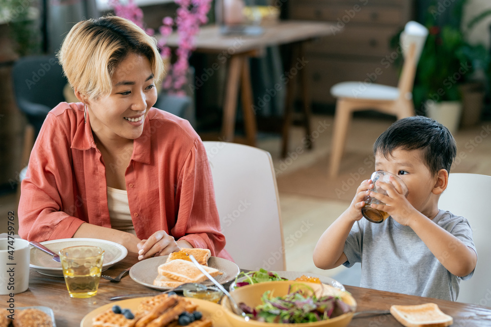 Happy young Asian woman looking at her adorable little son drinking tea or juice by breakfast