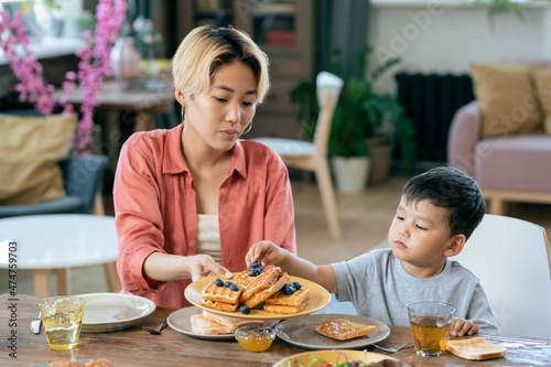 Young attractive woman passing her cute little son some waffles with blueberries during breakfast in home environment