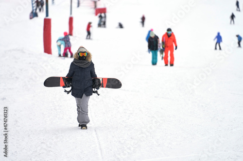  A snow-covered mountainside with a t-bar lift and tourists skiing and boarding. In the foreground is a girl with a snowboard walking up the slope. Selective focus.
