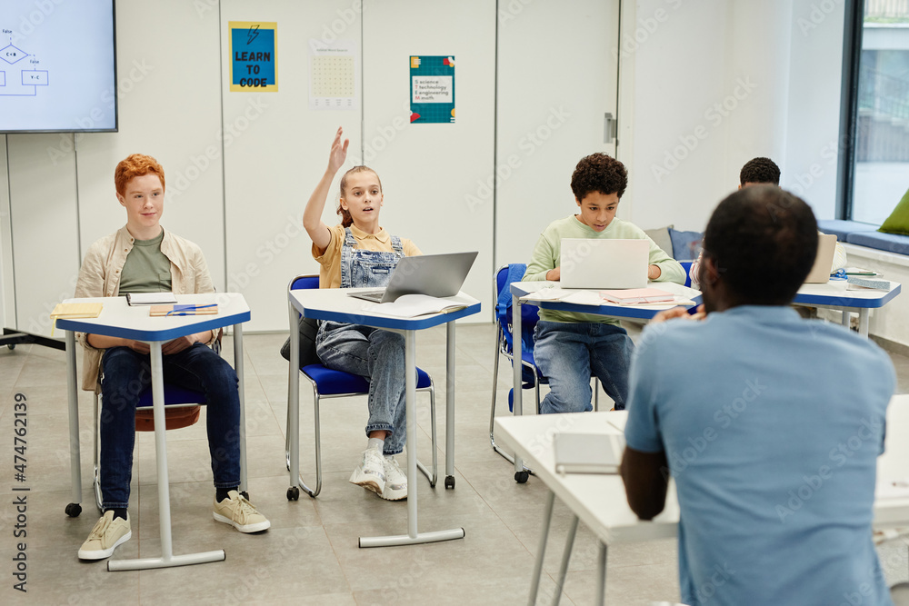 Obraz premium Diverse group of chilrden sitting at desks in modern school classroom focus on teenage girl raising hand, copy space