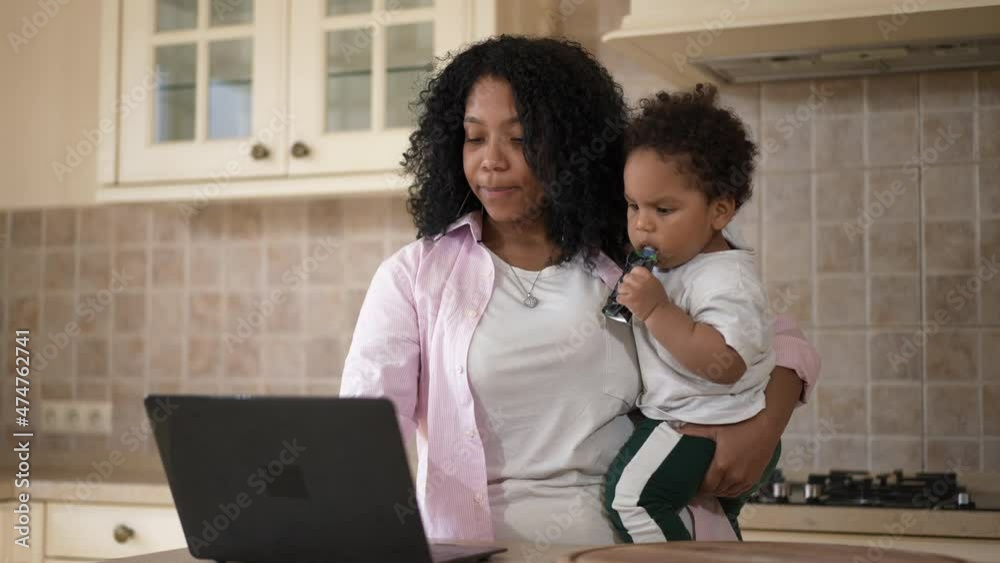 Young busy African American mother kissing toddler son messaging online on laptop. Positive beautiful woman with baby working remotely at home in kitchen. Freelance and family balance Young busy African American mother kissing toddler son messaging online on laptop. Positive beautiful woman with baby working remotely at home in kitchen. Freelance and family balance