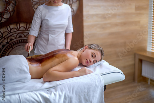 Spa therapy for young calm caucasian female receiving cosmetic mask on back spine at beauty salon, naked masseur applying mask with brush. beauty, wellness, healthy skin concept