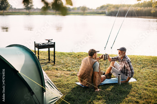 Happy senior man and his adult son toast while fishing on their camping day.