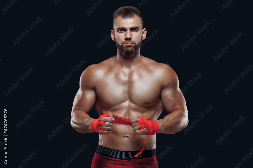 Muscular young fighter is applying bondage tape on hands getting ready to fight isolated on black background