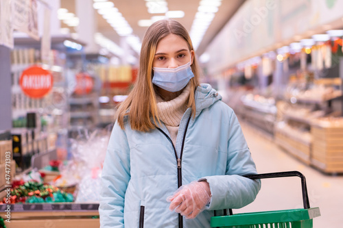 Front view of a young beautiful girl standing in a supermarket in a medical mask and gloves holding a shopping basket. Shopping during quarantine in compliance with the rules to prevent the spread of
