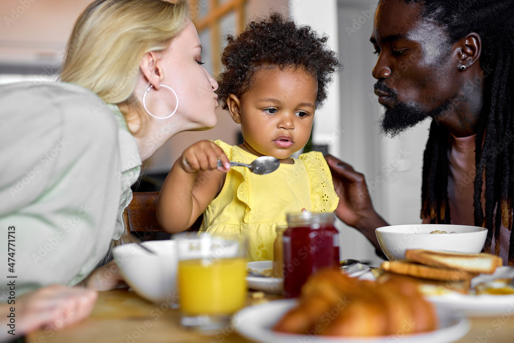 Cute black toddler girl enjoying Breakfast with parents at home, pretty child eating porridge. Healthy food for young children. interracial family at home at weekends, holidays, in the morning