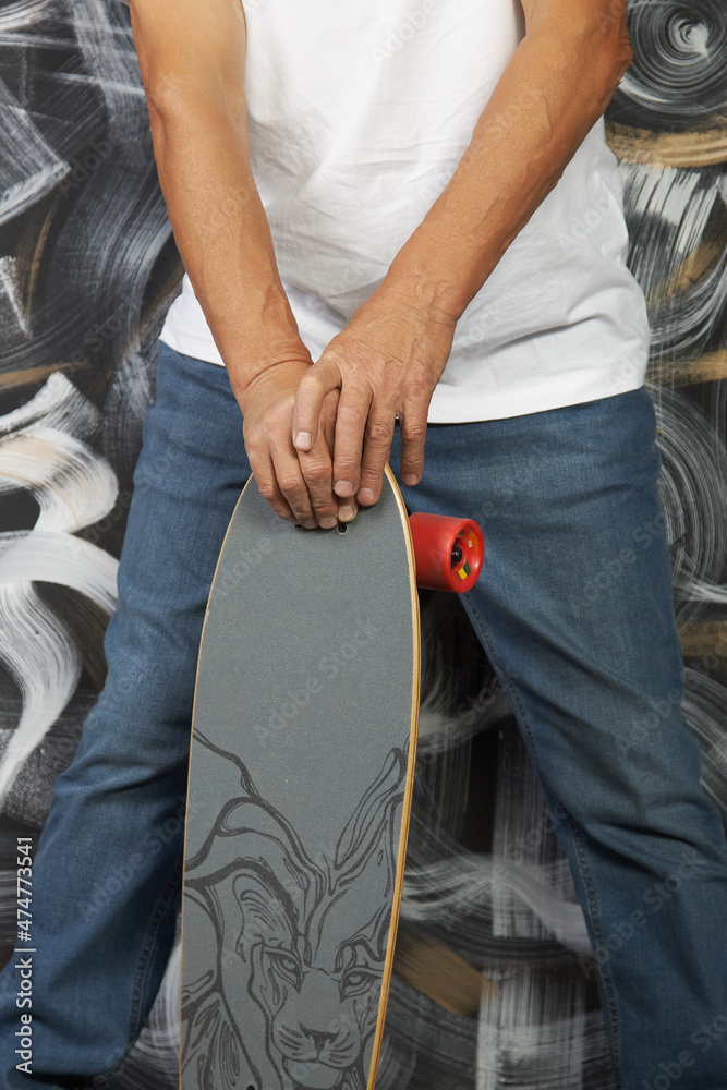 Legs of a fashionable man in jeans and sneakers with a skateboard in his hands close-up. A man poses against a painted wall. Vertical photography