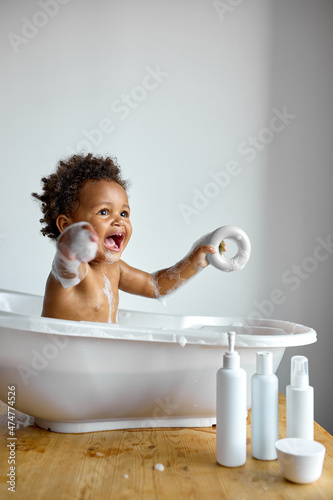 Obraz na plátně Happy little black baby girl sitting in bath tub playing with toys in bathroom