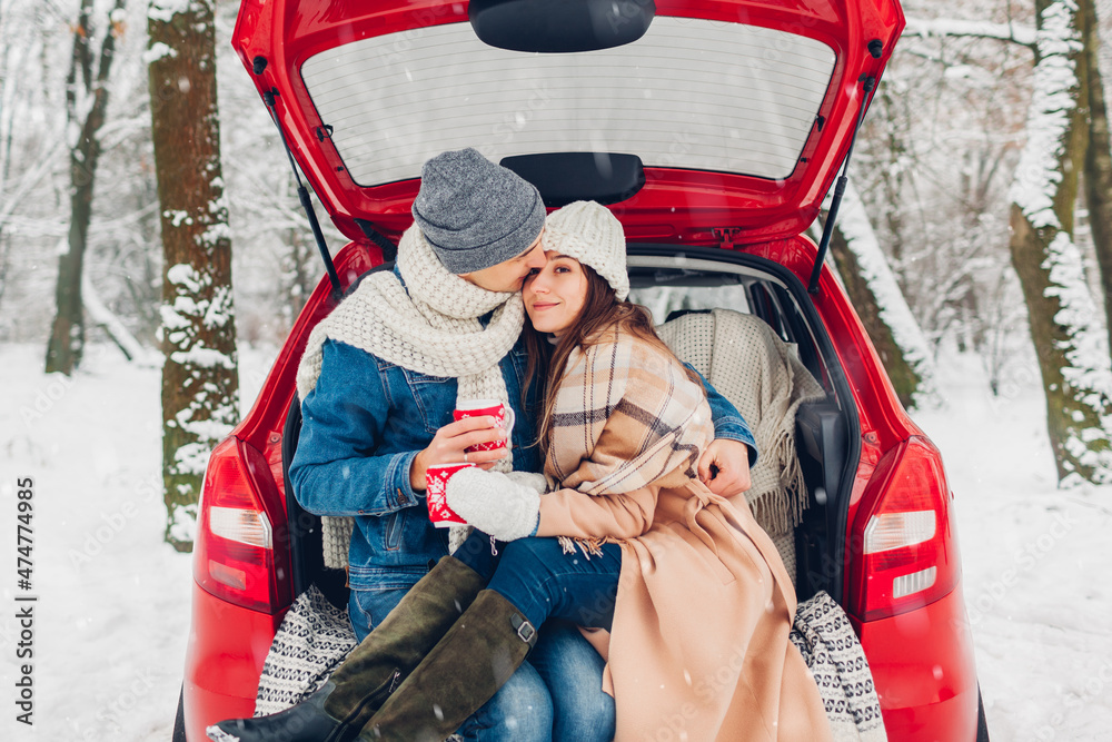 Couple in love sitting in car trunk drinking hot tea in snowy winter forest. People relaxing outdoors during road trip