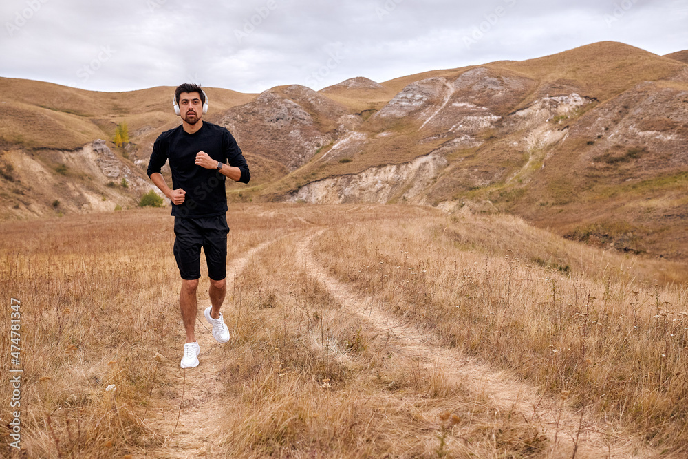 outdoor image of young fit runner man running in the morning in mountains. Fitness male exercising before sunrise and listenting the music on white headphones earphones. People and sport concept