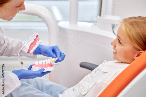 Dental consultation. Woman dentist holds model of human jaw in hands. Stomatologist telling about Oral hygiene to child girl patient sitting on couch. Taking care of dental health. Medicine concept