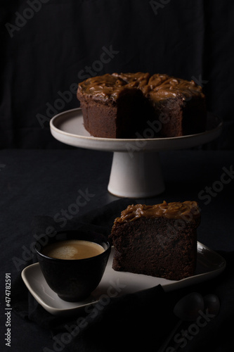 A big chocolate cake slice and a cup of coffee over a ceramic tray with a blurred cake as background.