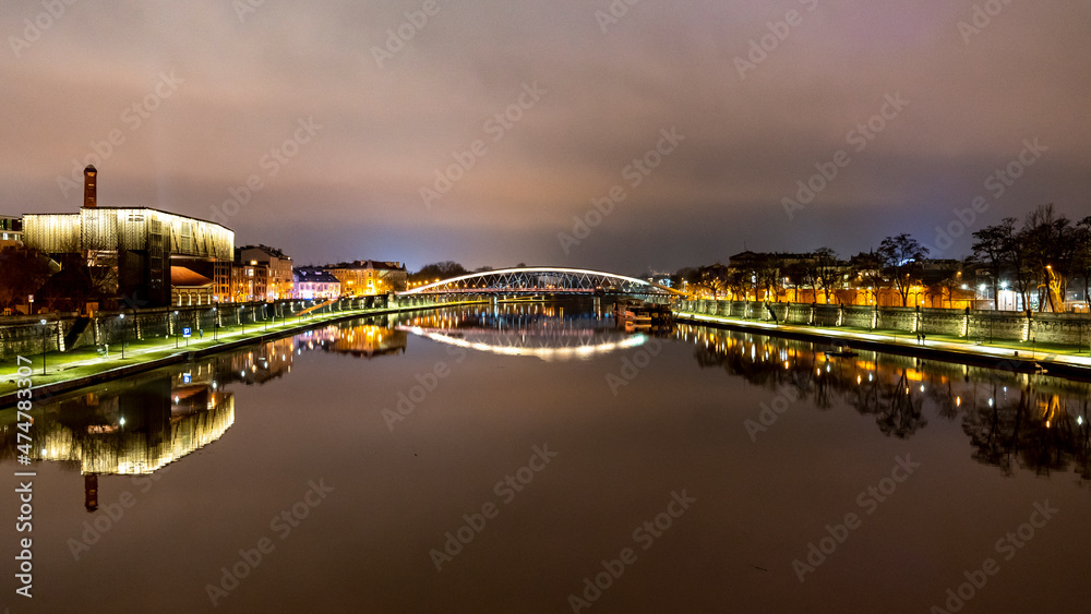 Wieczorne ujecie rzeki Wisła w centrum Krakowa z oświetlonymi budynkami po jej brzegach - obrazy, fototapety, plakaty 