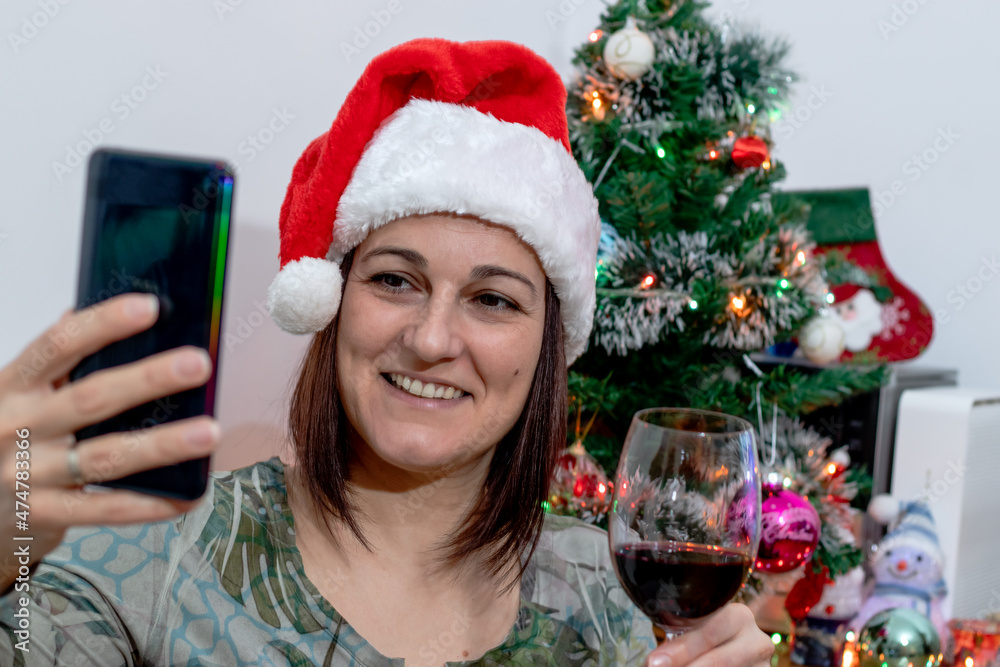 Woman with Santa Claus hat, in front of Christmas tree, holding a glass of wine and taking a selfie or talking to her family via a video call on her smartphone. Being isolated for holidays.