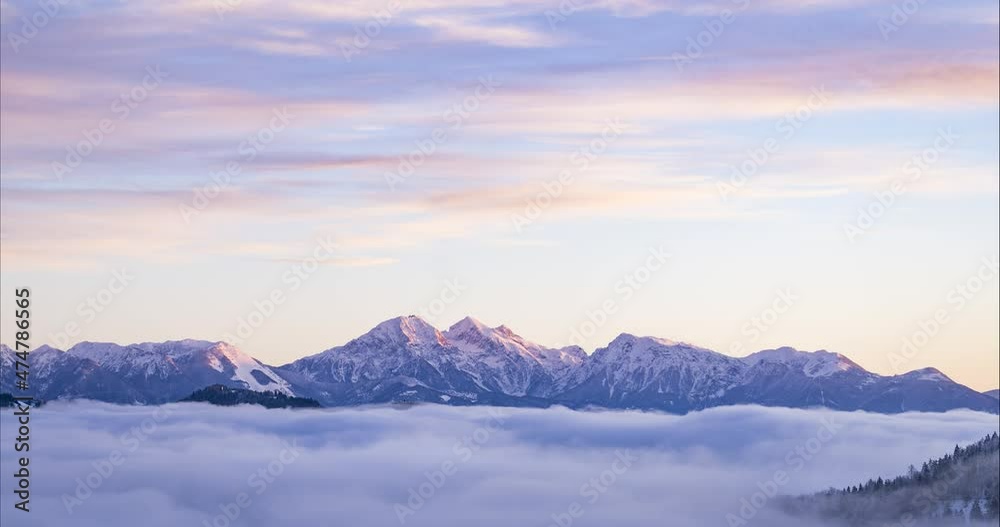 Low inversion clouds moving in alpine valley. Time lapse of fog covering amazing landscape in winter. Snowy mountains in the distance. Pristine nature in Slovenia, Europe. Sunrise. Static shot
