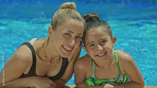 Mother and daughter are resting by the pool. Daughter and mother chatting in the pool, spending time together, relaxing by the pool. They are smiling looking at the camera. Happy family on vacation.