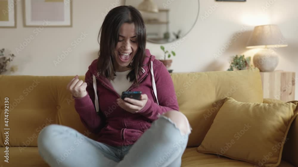Joyful woman reading good news in unexpected message or email on mobile phone in apartment. Surprised young female checking phone and celebrating while sitting on couch at home. 