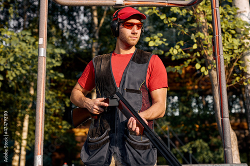 Young caucasian man skeet shooting outdoors. shooting clay pigeon targets in outdoor range, stands looking at side in contemplation, wearing protective safety goggles and headset, in cap photo