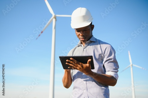 African engineer wearing white hard hat standing with digital tablet against wind turbine on sunny day photo