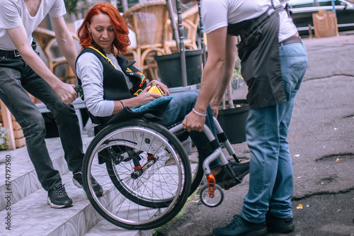 Waiters lift a girl in a wheelchair up the steps without a ramp. A woman is going to have lunch in a cafe inaccessible to people with special needs