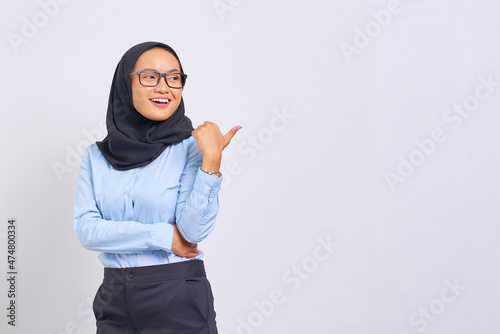 Portrait of smiling young Asian woman pointing thumbs at copy space isolated on white background