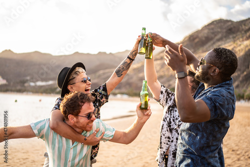 Group of happy young multi ethnic people having fun together on the beach.  Smiling friends raising a toast with beer.  Summer sunset time. Multiracial girls and guys drinking, photo