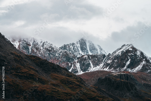 Atmospheric alpine landscape with great mountain peaked top with snow in low clouds. Dramatic mountain scenery with sharp pinnacle in overcast weather. Awesome view to snowy pointy peak in cloudy sky.