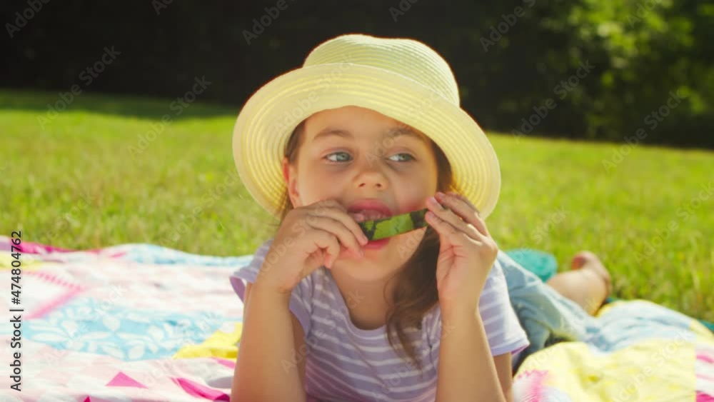 Preschool girl in a headdress has a watermelon in the park in the summer on a picnic. Cute little girl is eating a juicy watermelon lying on the grass in a clearing in the forest on a sunny summer day Preschool girl in a headdress has a watermelon in the park in the summer on a picnic. Cute little girl is eating a juicy watermelon lying on the grass in a clearing in the forest on a sunny summer day