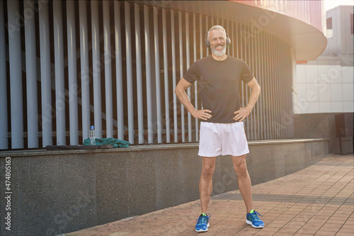 Full length shot of joyful sportive middle aged man in sportswear and headphones smiling while warming up, getting ready for workout outdoors