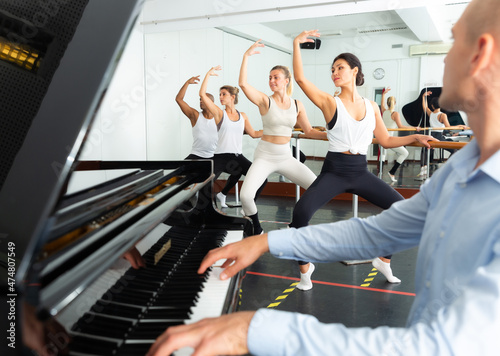 Ballet students doing exercises near barre with musician at piano in foreground