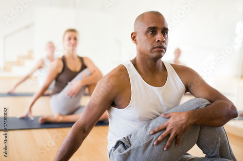 Focused hispanic man doing yoga with group of people in fitness studio, sitting in twisting asana Matsyendrasana
