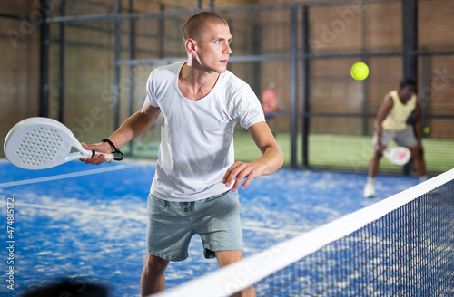 Portrait of concentrated paddle tennis player preparing to hit forehand to return ball on indoor court.. © JackF