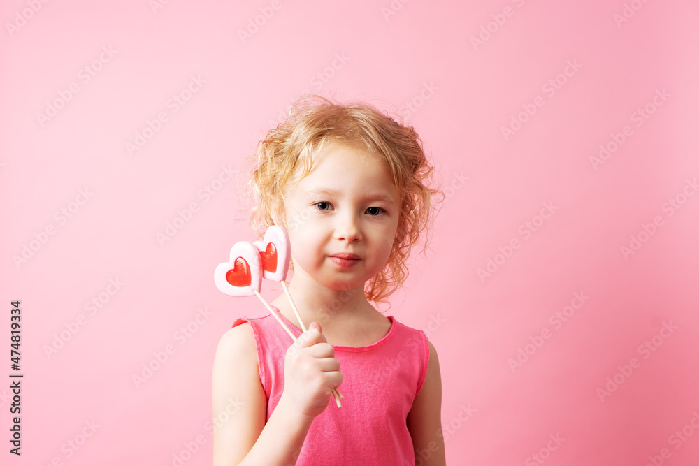 Little girl holding large heart-shaped lollipops in her hands on a pink background.