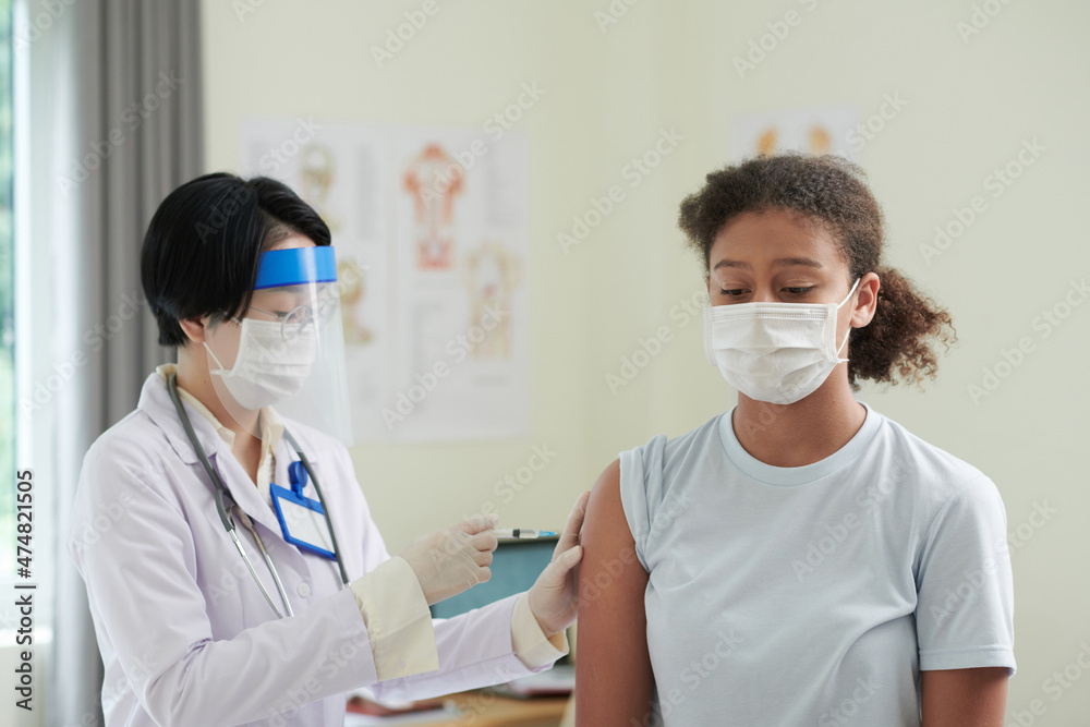 Serious teenage girl in medical mask getting covid-19 vaccine at hospital