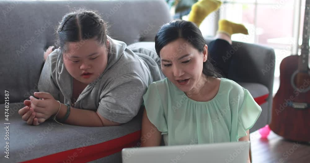 Delighted daughter with down syndrome and loving Asian mother using laptop and entertaining together in living room at home Delighted daughter with down syndrome and loving Asian mother using laptop and entertaining together in living room at home