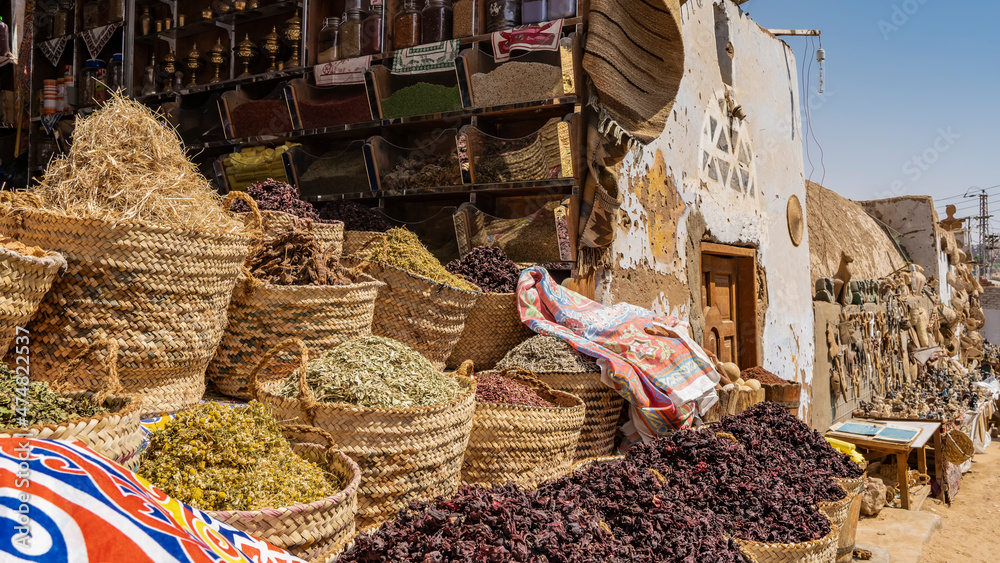 Various dried herbs, spices, and karkade tea are sold at the Egyptian street market. Goods in large wicker baskets and on shelves. The wall of the house with peeling whitewash is visible.