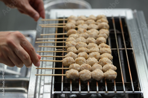 A merchant is flipping a skewer of pork meatballs with his hand to cook it on a smokeless electric grill. (selective focus)