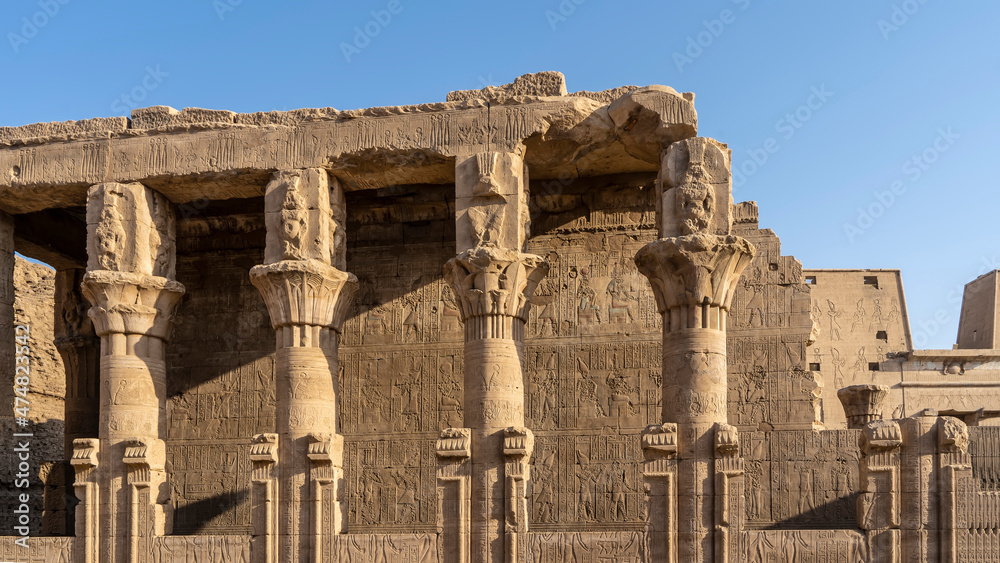 The dilapidated colonnade and wall in the ancient temple of Horus in Edfu are decorated with hieroglyphs, carved drawings. Capitals of columns of various shapes. Clear blue sky. A sunny day. Egypt