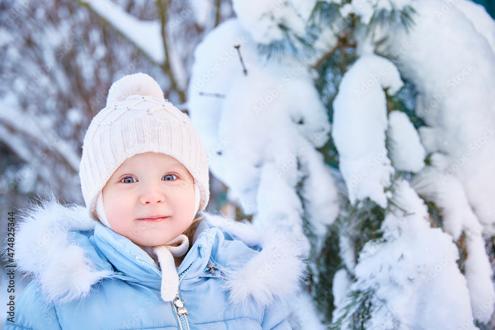 Little toddler boy in blue jacket and knitted hat standing in a snowy forest or park on a sunny day, smailing and looking at the camera Little toddler boy in blue jacket and knitted hat standing in a snowy forest or park on a sunny day, smailing and looking at the camera