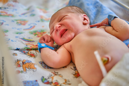 A newborn baby with a maternity hospital bracelet on his arm is crying in a crib. A newly born child in a clinic bed behind a transparent glass