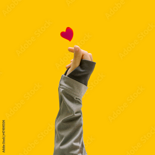 K pop concept. A girl teenager hand showing finger heart gesture. Red heart above. Unique yellow background.