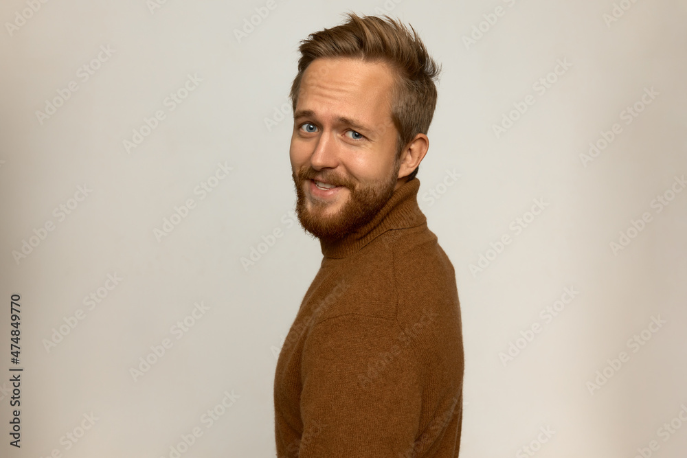 Obraz premium Handsome photogenic adult guy in good mood wearing cashmere turtleneck with stylish beard and haircut standing half-turned to camera, smiling against white wall with copy space for your content