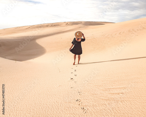 Mulher de vestido e chapéu caminhando nas dunas de areia  © carina furlanetto